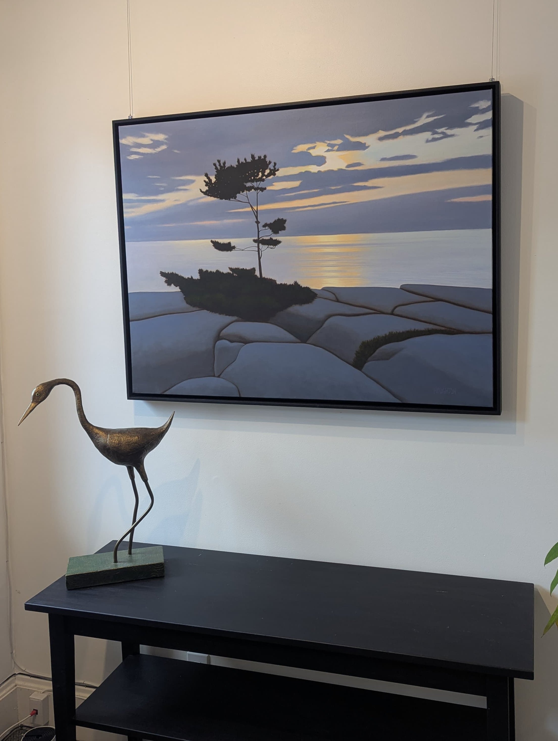 Framed artwork of a sunset over water with a crane on a black table in a room.